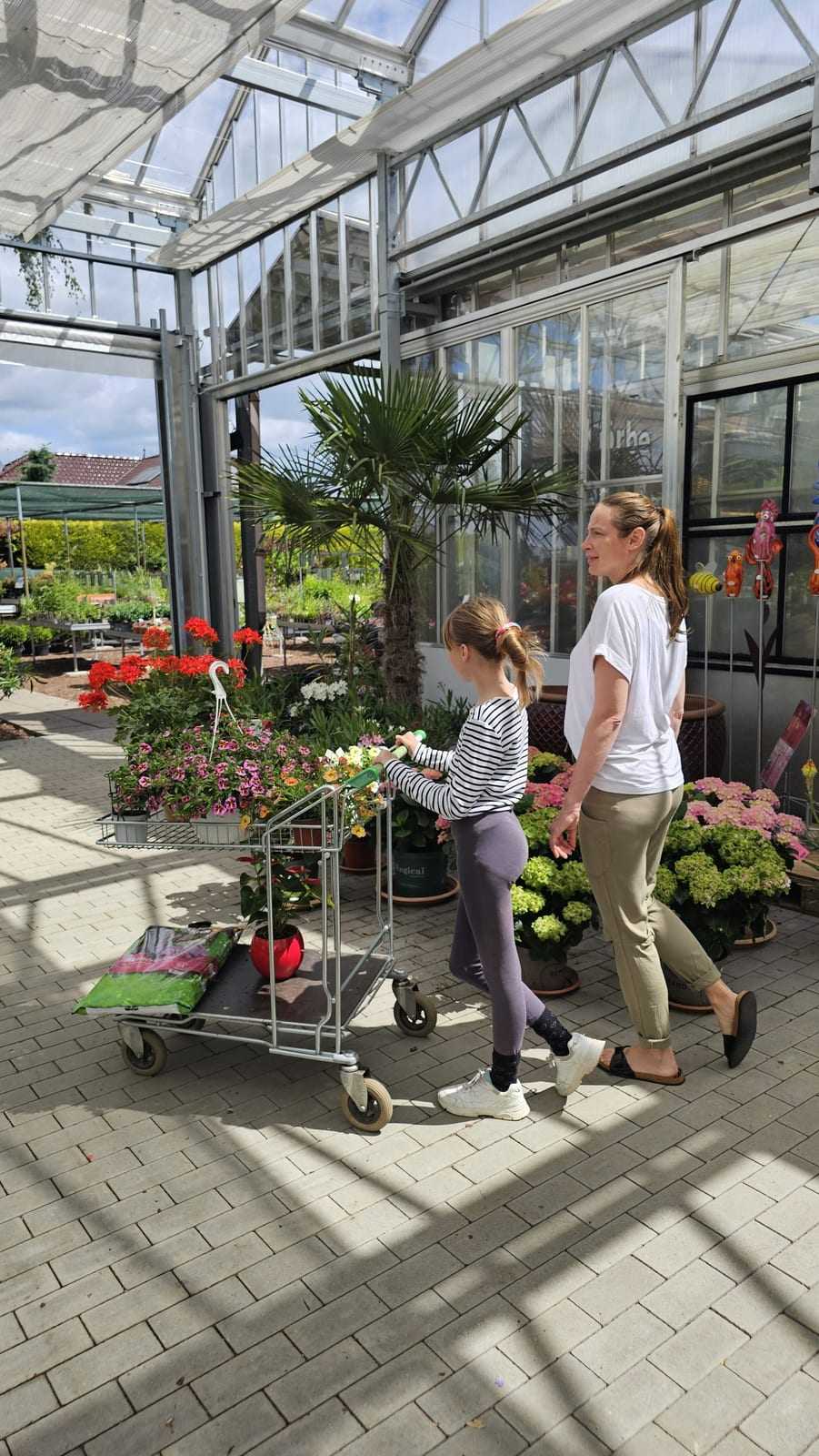 Mutter und Tochter ziehen einen bepflanzten Wagen durch den sonnigen Innenhof der Baumschule
