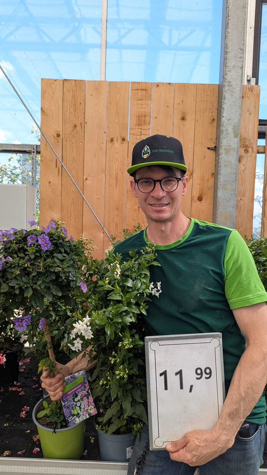 Mitarbeiter mit Kappe hält lachend eine blühende Pflanze und ein Preisschild vor einer Holzwand
